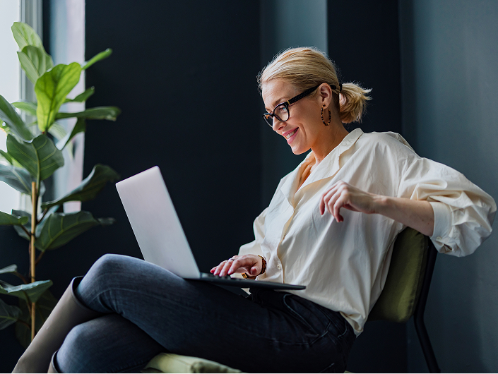side view of a woman sitting on a chair, one arm on the back, with a laptop on her lap